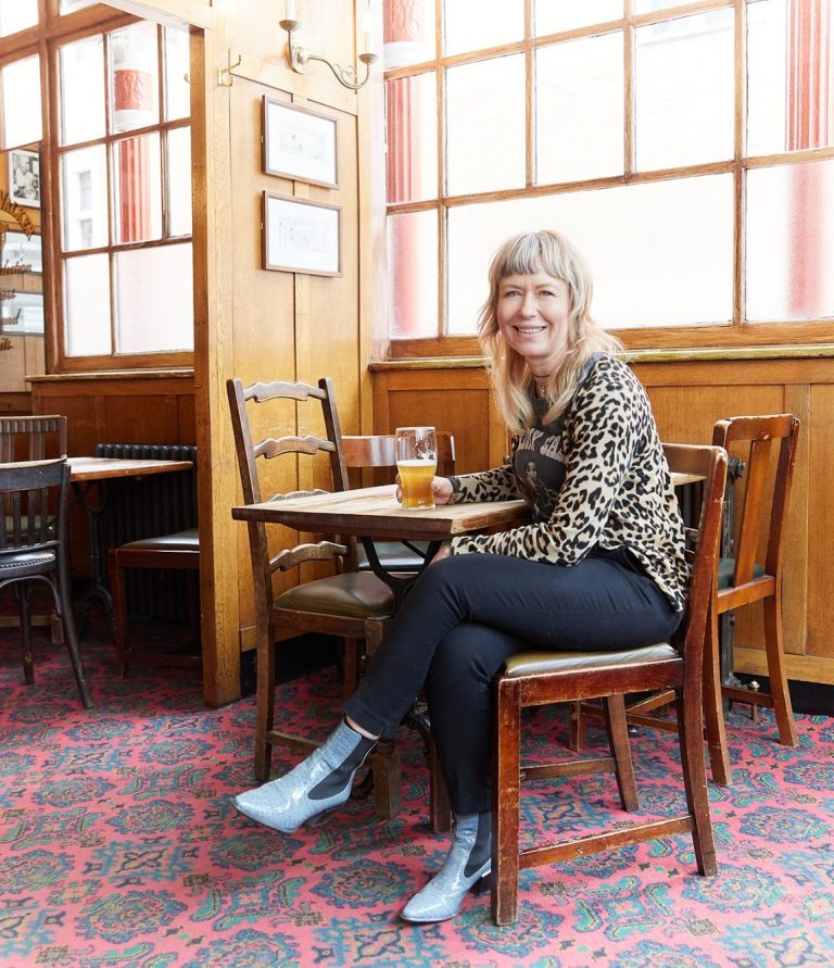 Shaz portrait Sharon wears a leopard print top, black jeans and blue boots, sitting at a table in a pub, smiling and holding a drink.