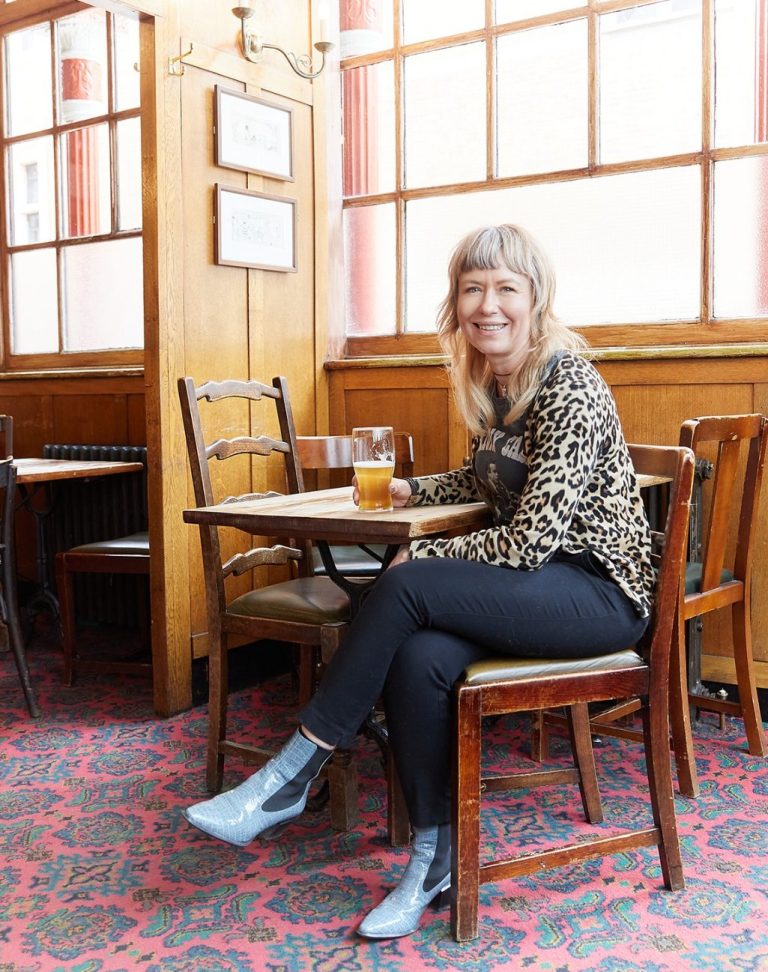 Sharon wears a leopard print top, black jeans and blue boots, sitting at a table in a pub, smiling and holding a drink.